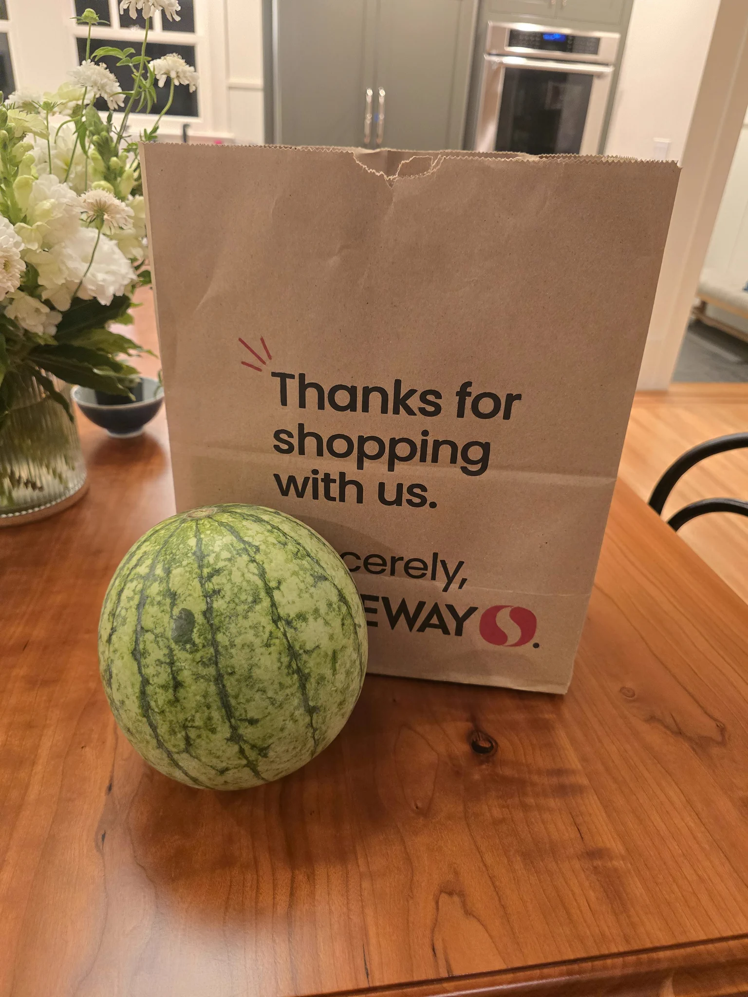 A whole round watermelon on a wooden kitchen table, with a brown paper shopping bag visible in the background